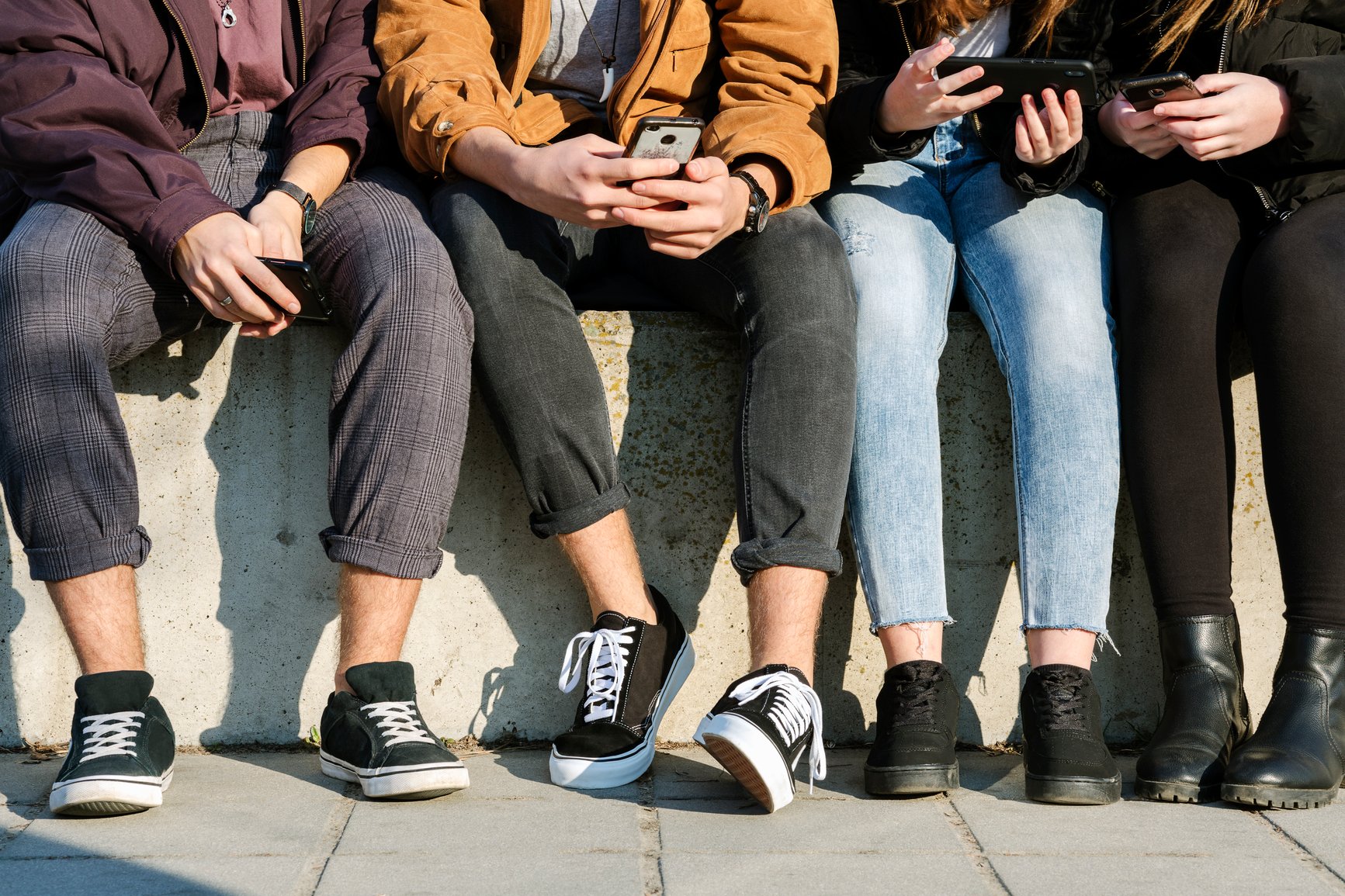 Anonymous group of teenage generation z friends checking their mobile phones while sitting together.